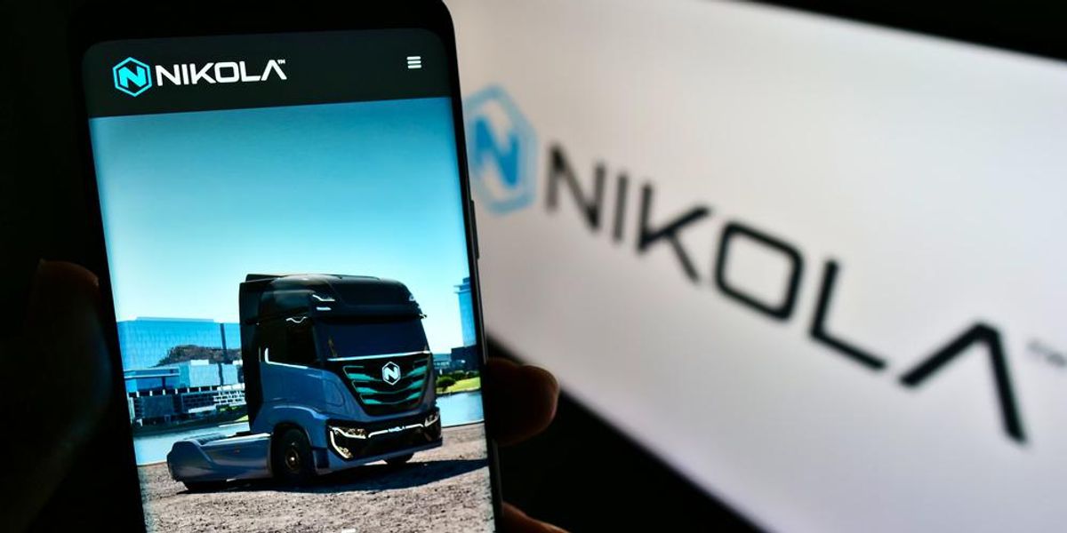 A Nikola elektromos kamiongyártó vállalat applikációja egy telefonon, amit egy ember tart a kezében, a kijelzőn egy kék kamion látható, a kép hátterében elmosódottan a Nikola logója látható