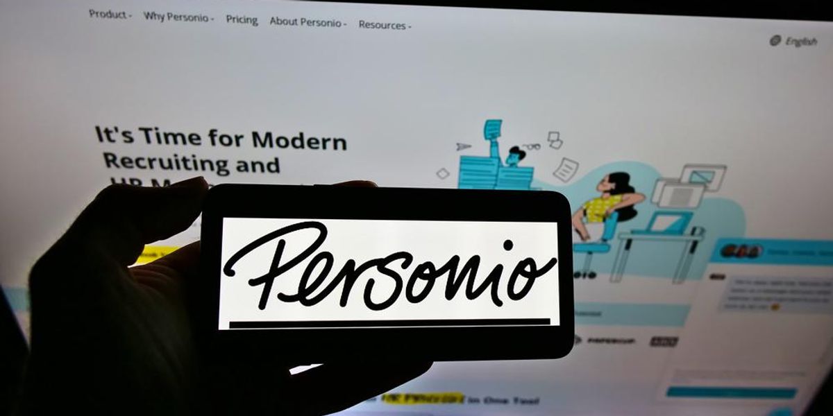 A Personio HR-szoftvercég logója egy telefonon, amit egy ember tart a kezében, a háttérben a Personio weboldala látható