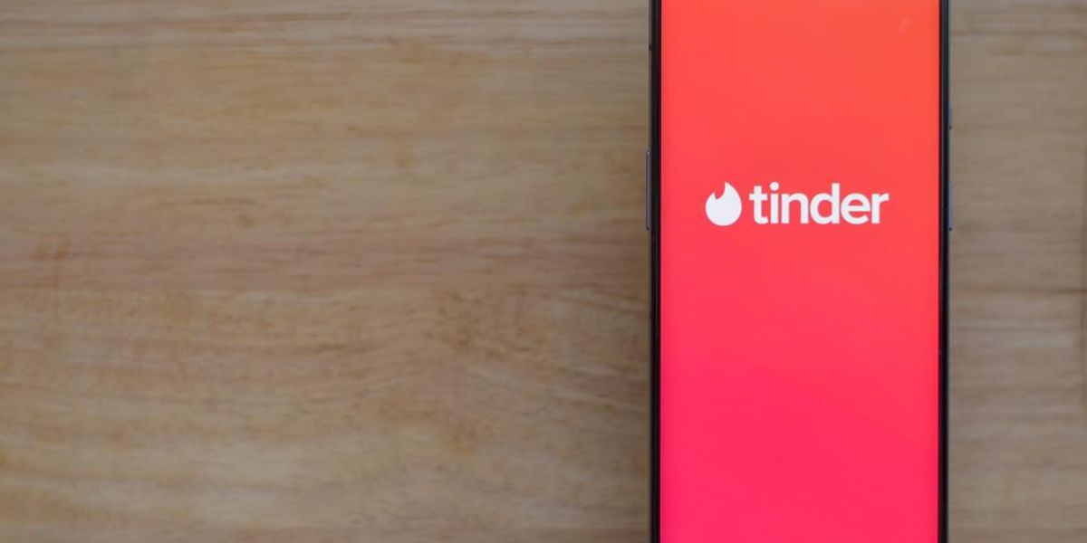 A Tinder randiapp fehér logója egy narancssárgás-lilás háttérben egy okostelefon képernyőjén, ami egy fa felületen van