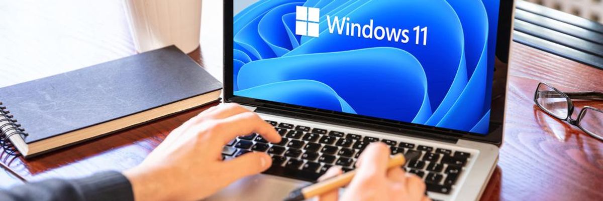 A Windows 11 operációs rendszer tölt egy szürke laptopon, ami egy ember használ, akinek egy toll van a kezébe, a gép egy faasztalon van, amin egy jegyzetfüzet is található