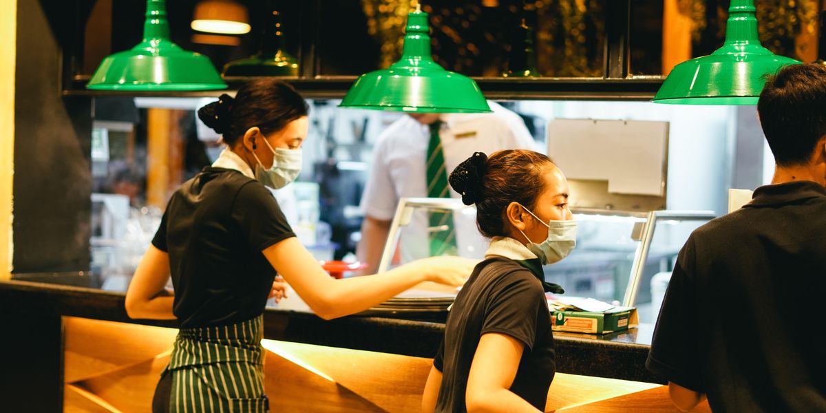 Arcmaszkos, fekete felsős, zöld-fehér csíkos kötényes pincérnők szolgálják fel az ételeket a vendégeknek egy étteremben