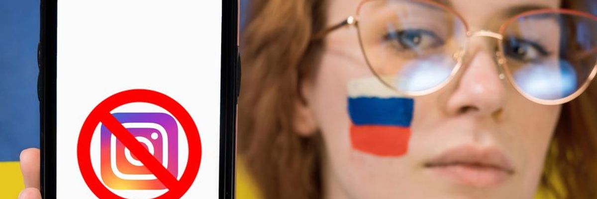 Az Instagram áthúzott logója egy telefon képernyőjén, amelyet egy szemüveges lány tart a kezében, akinek az orosz zászló van az arcára festve
