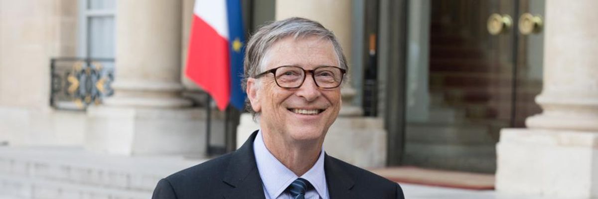 Bill Gates fekete zakóban, fehér ingben, kék-fehér-fekete nyakkendőben, szemüvegben mosolyog egy ablakos épület előtt