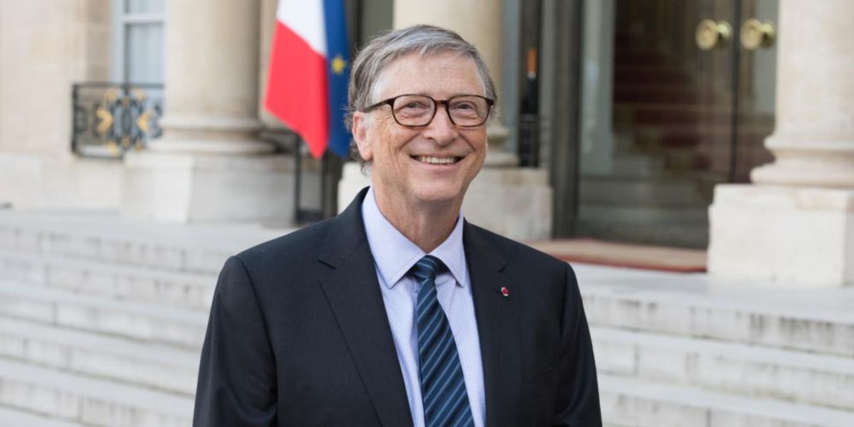 Bill Gates fekete zakóban, kék ingben és csíkos nyakkendőben vigyorog egy épület lépcsője előtt