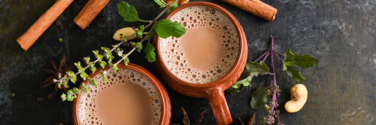 Chai teák barna bögrékben, körülöttük fahéj és egyéb fűszerek.