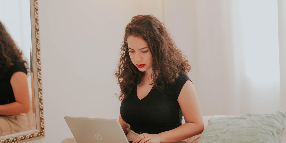 Dekoratív lány világos nadrágban, fekete felsőben ül az ágyán home officeban ölében egy laptoppal és dolgozik, mellette a falon egy tükör van