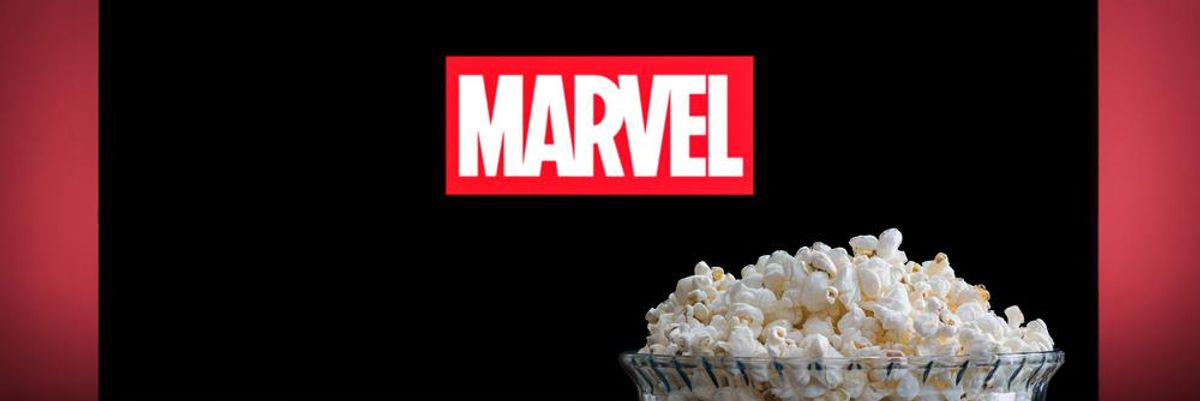 Egy adag pattogatott kukorica egy távirányító mellett és egy TV előtt, melynek lépernyőén a Marvel logója látható