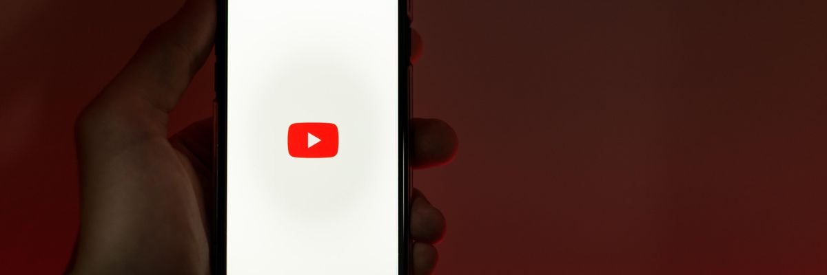 Egy ember egy okostelefont tart a kezében,a melynek képernyőjén a YouTube logója látható. A háttér piros-fekete.