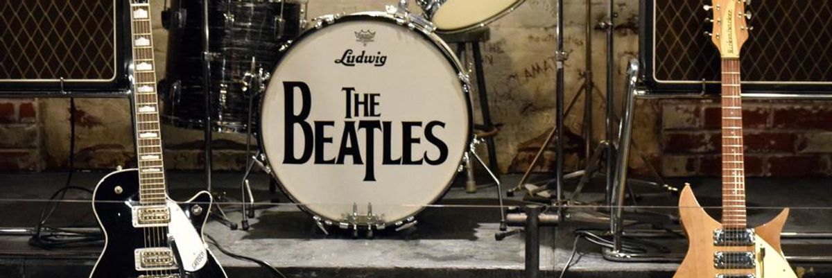 Egy fekete és egy barna elektromos gitár a színpadon, köztük egy emelvényen egy dobfelszerelés látható, a lábdobon a The Beatles logóval