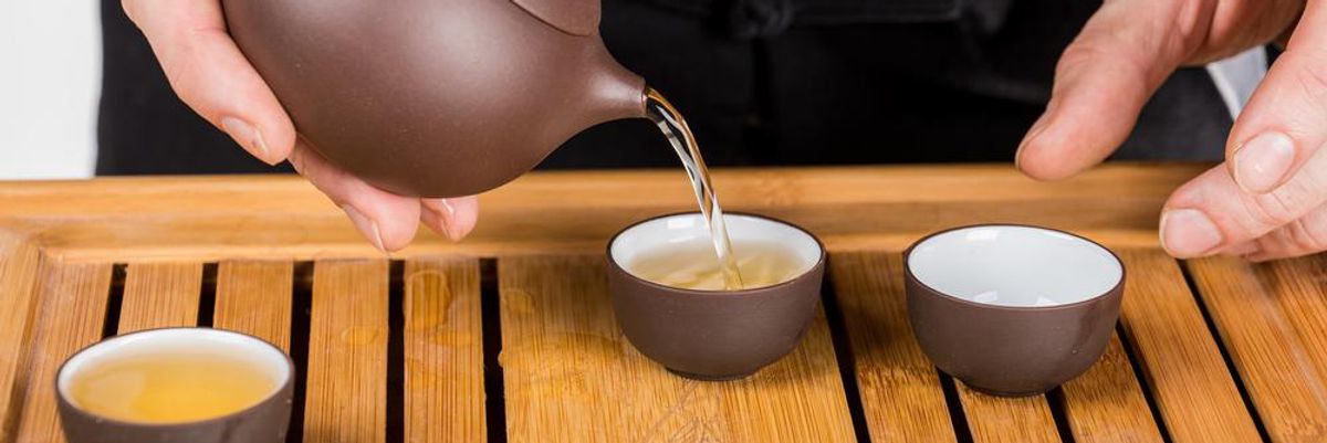 Egy fekete ruhás férfi épp teát önt egy zisha teáskannából három csészébe, amelyek egy fa tálcán vannak