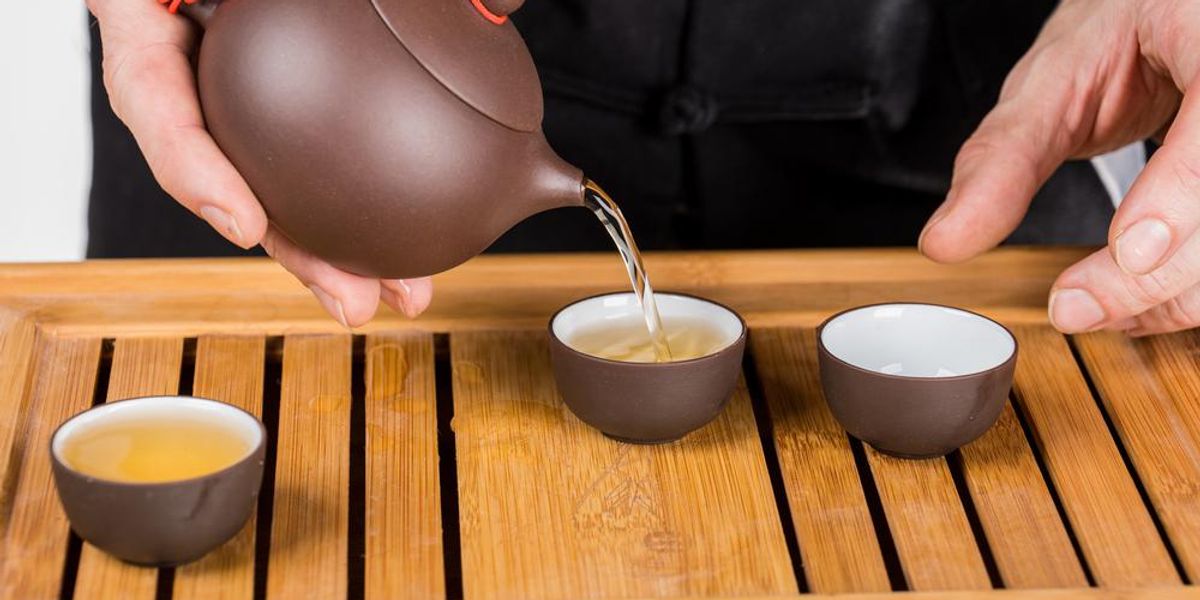 Egy fekete ruhás férfi épp teát önt egy zisha teáskannából három csészébe, amelyek egy fa tálcán vannak