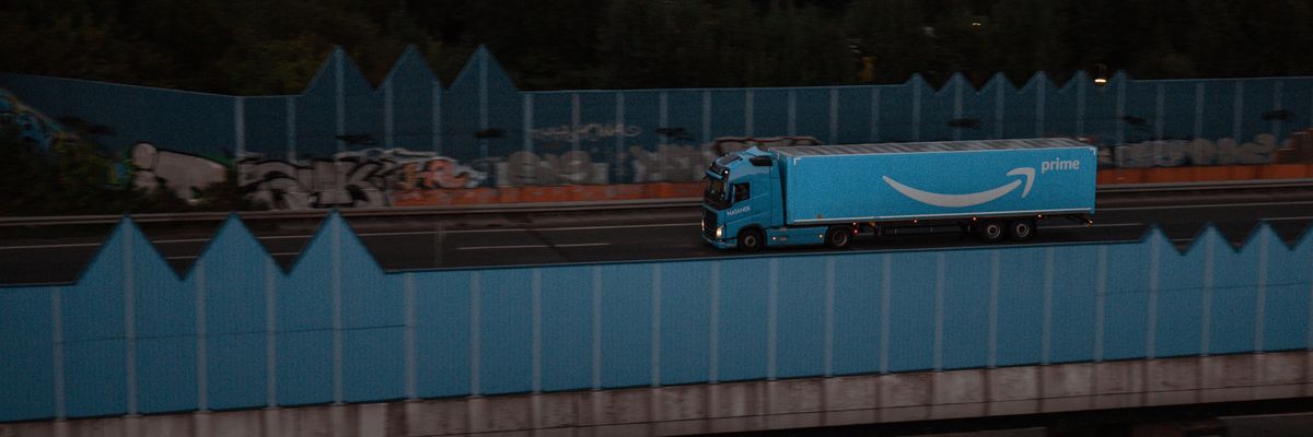 Egy kék színű Amazon Prime feliratos kamion halad át egy kék színű összefirkált fallal védett hídon esti szürkületben