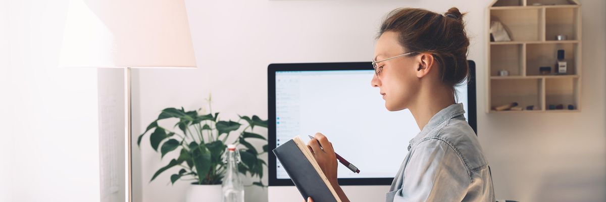 egy nő ír a noteszébe a számítógép képernyője előtt ülve