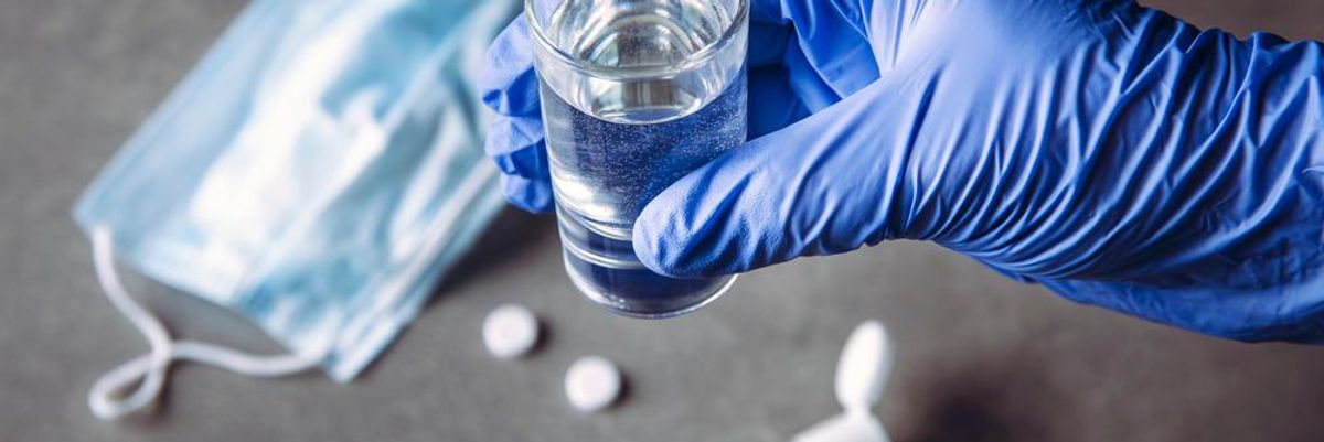 Egy pohárnyi víz fertőtlenítő szerrel vegyítve, egy orvosi kesztyűt viselő ember tartja a poharat, a háttérben maszk, tabletták és egy kézfertőtlenítő