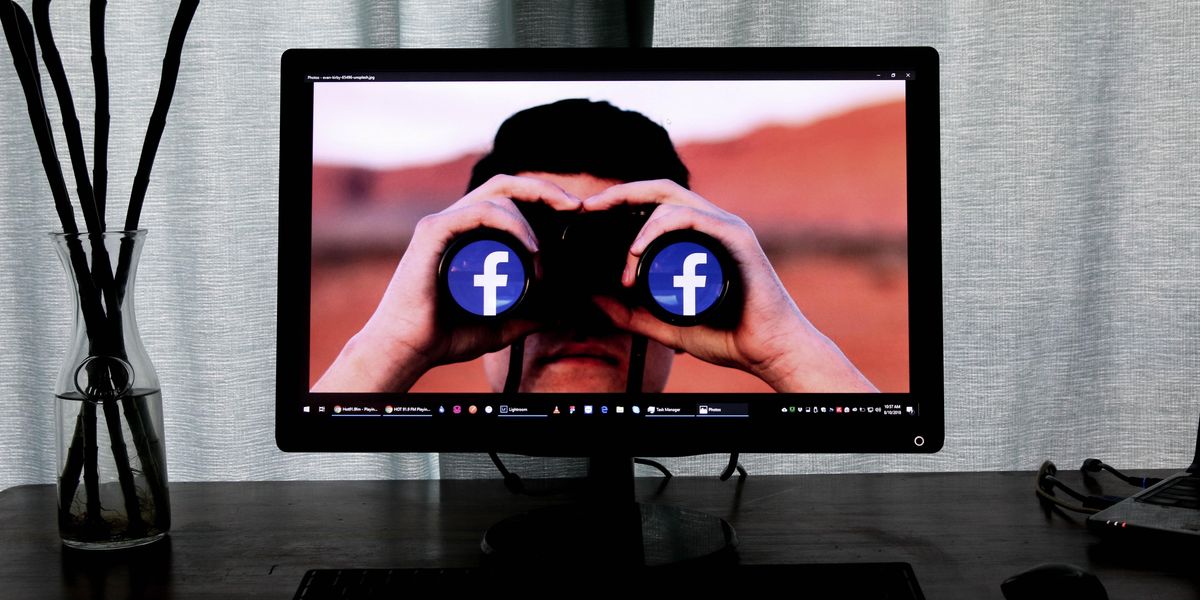 Egy sötét asztalon van egy egér, egy klaviatúra, egy váza és egy monitor, a monitoron egy ember látható, aki távcsővel néz, a távcső lencséiben a Facecbook logója van