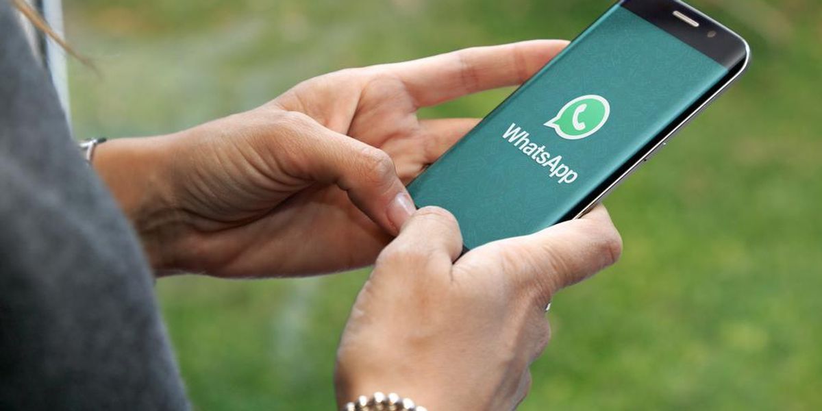 Egy szürke felsős, karkötős nő a kezében tartja okostelefonját, amin a WhatsApp alkalmazása töltődik, a háttérben egy zöld füves terület látható