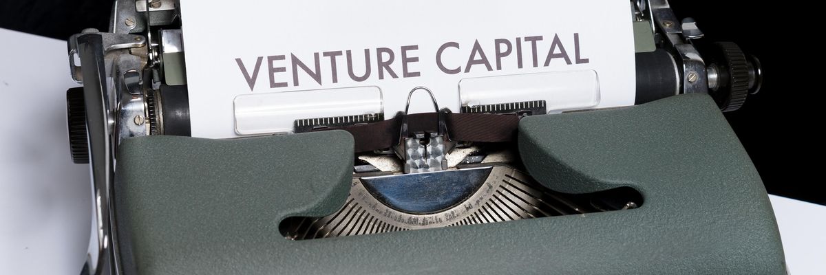 Egy szürkés-zöldes írógépen a Venture Capital szavakat gépelték be, ami kockázati tőkét jelent, az írógép egy fehér asztalon van, a háttér sötét