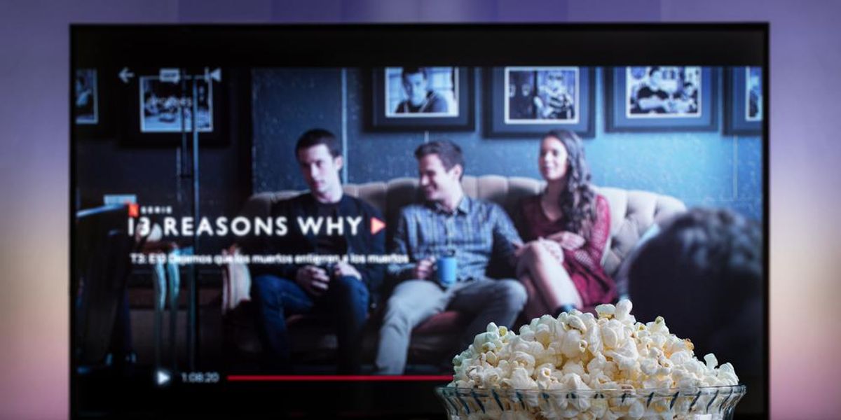 Egy tál popcorn és egy távirányító látható egy fa felületetn egy TV előtt, amelynek képernyőjén a Netflix 13 okom volt című sorozata megy
