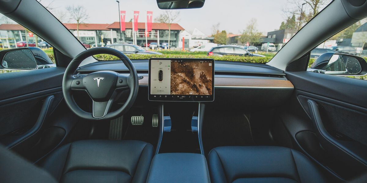 Egy Tesla belseje fekete bőr ülésekkel és fedélzeti számítógéppel, amelynek képernyőjén az autó és a környezet látható. Az autó a Tesla márkakereskedése előtt áll.