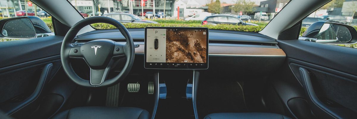 Egy Tesla belseje fekete bőr ülésekkel és fedélzeti számítógéppel, amelynek képernyőjén az autó és a környezet látható. Az autó a Tesla márkakereskedése előtt áll.