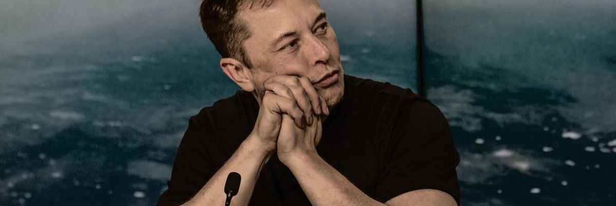 Elon Musk fekete pólóban könyököl  egy asztalon egy mikrofon előtt