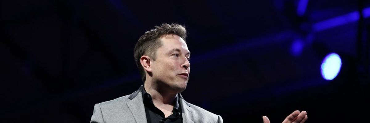 Elon Musk szürke zakóban és fekete ingben a kezét felemelve beszél egy színpadon