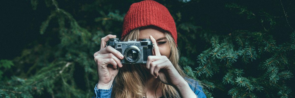 Farmerkabáros, piros sapkás szőke lány fényképezőgépet tart a kezében fenyőfák előtt
