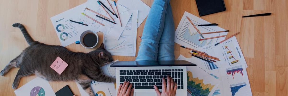 Home officeban dolgozó munkavállaló a padlón ül, ölében egy laptoppal, körülötte cetlik, papírok és egy macska van