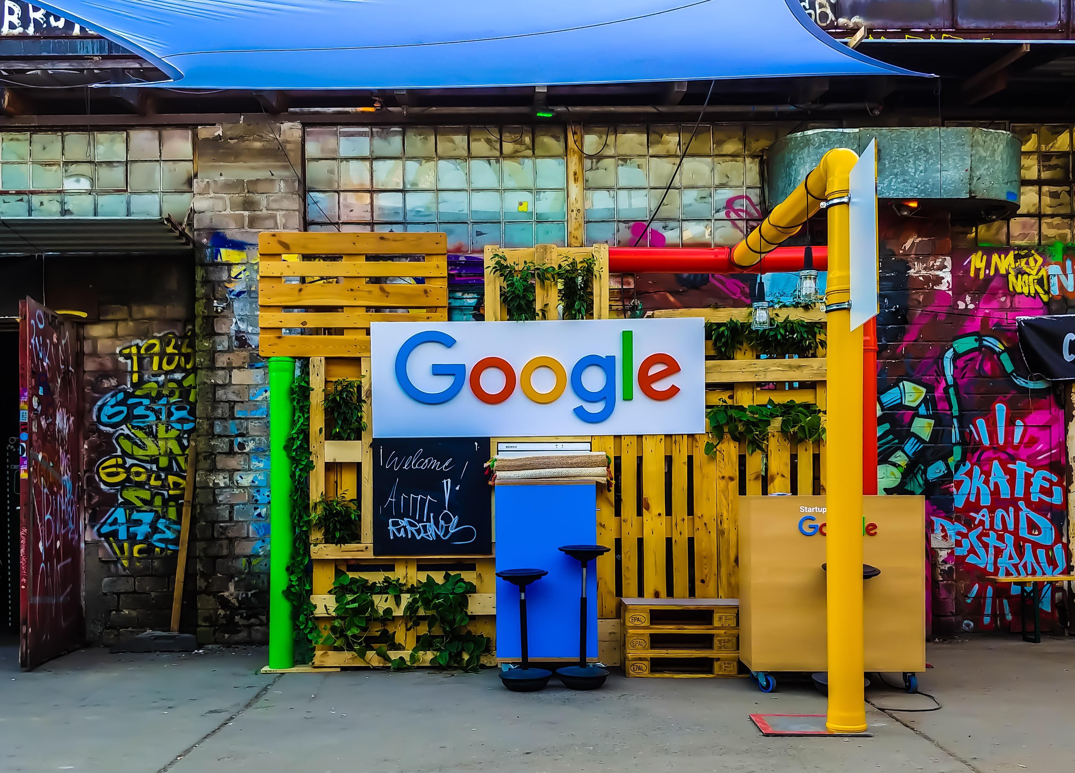 A Google logója egy sárga raklapokból összeállított installáción egy összegraffitizett fal előtt