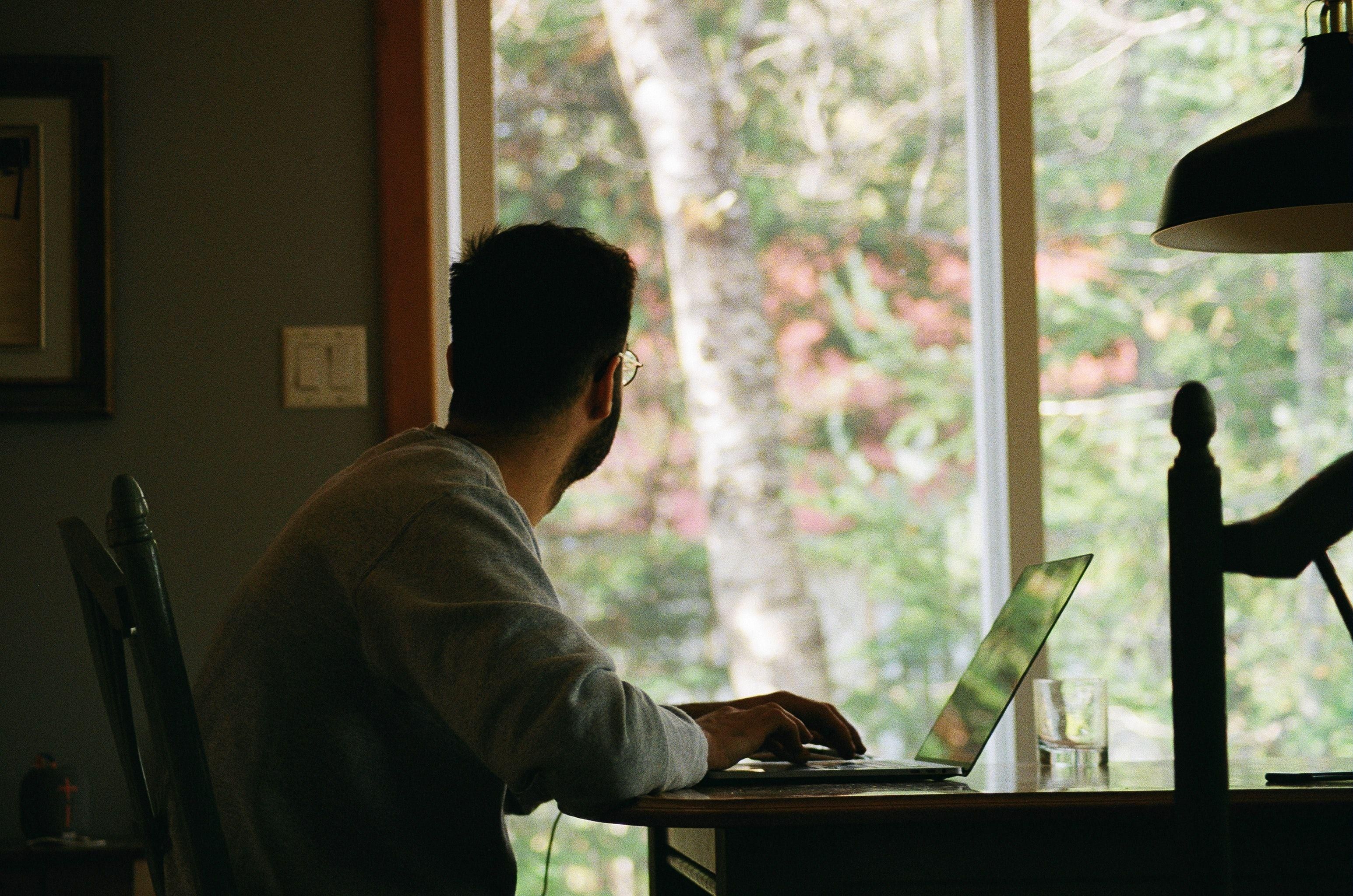 Ablakon kinéző otthon laptopján dolgozó férfi