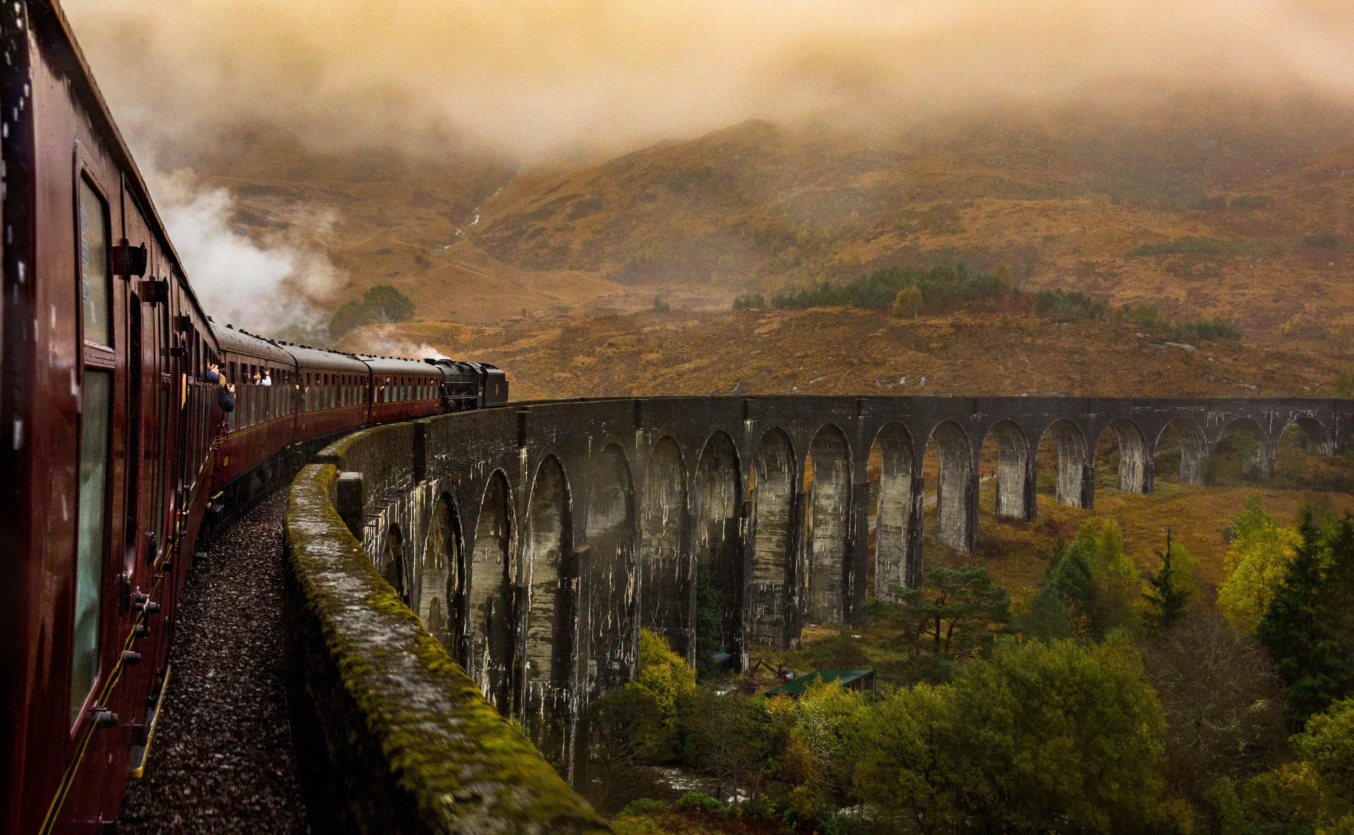 Piros vonat halad át egy hídon dombok között füstölve