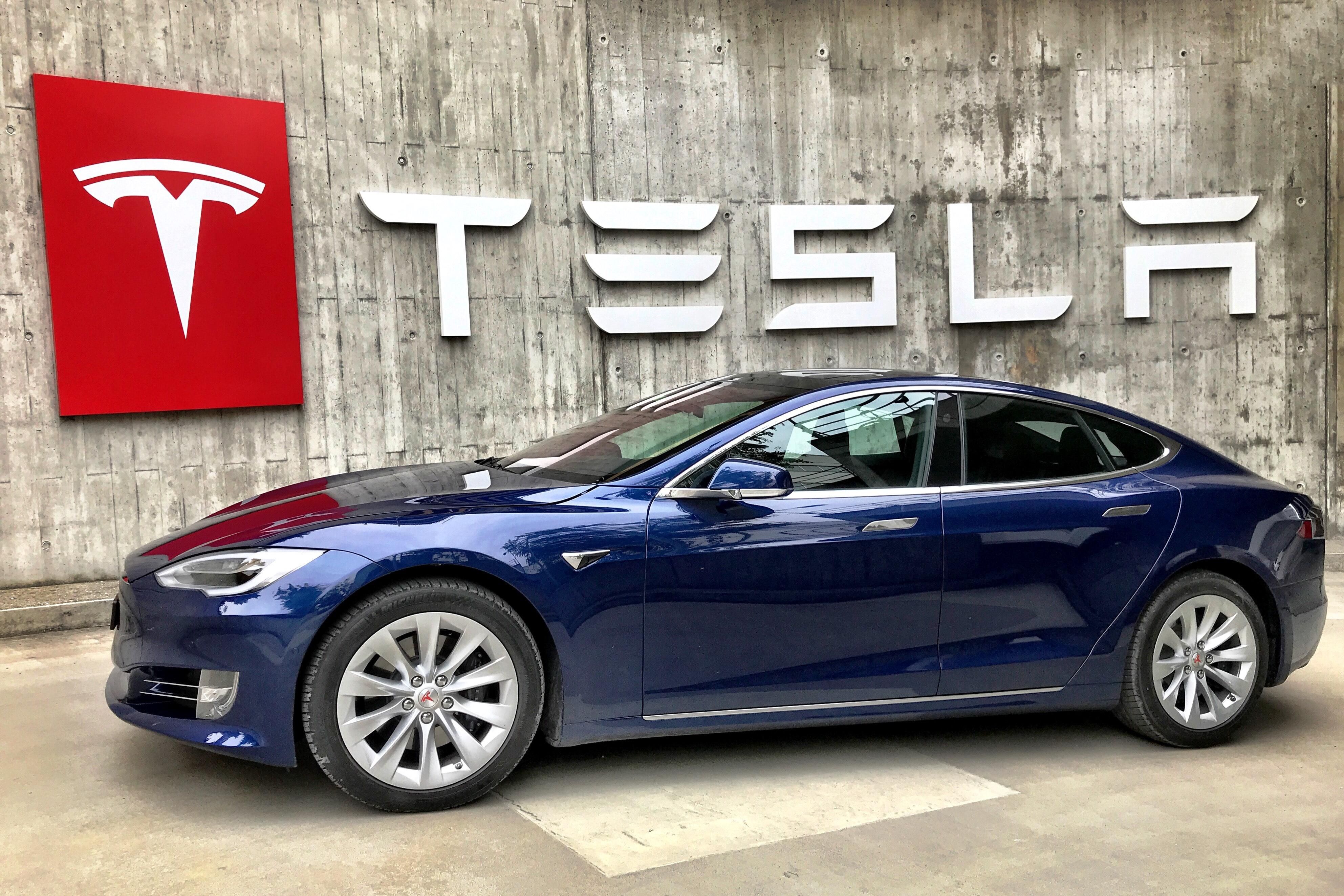 Kék Tesla jármű a Tesla logója és felirata előtt