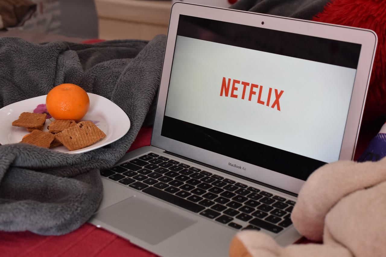 Netflixet néz valaki egy laptopon, ami az ágyon van, mellette egy tányér, amiben narancs és kekszek vannak