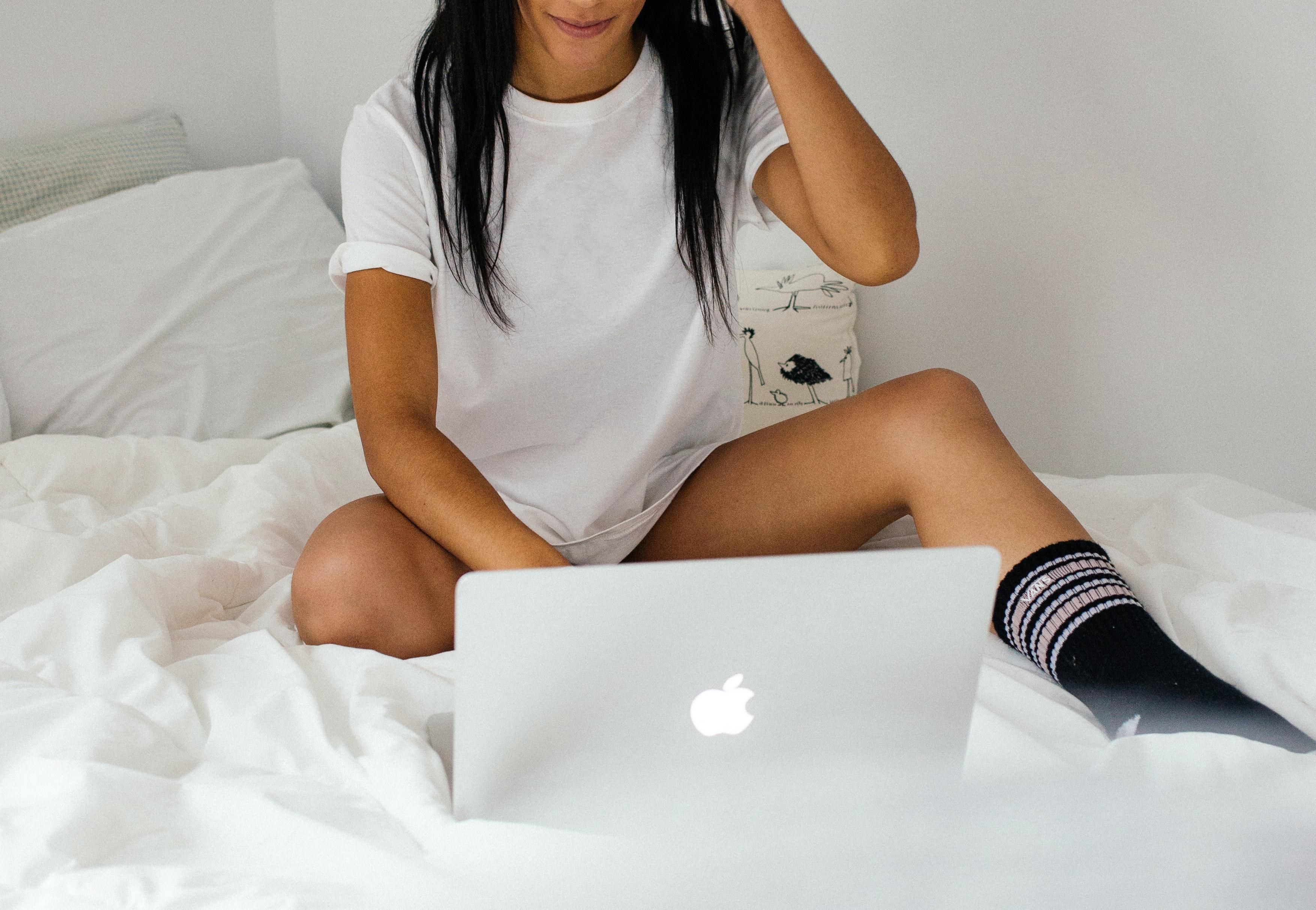 Szép, fekete hajú, fehér pólós, fekete zoknis lány épp OnlyFans tartalmat tölt fel a MacBook-ján