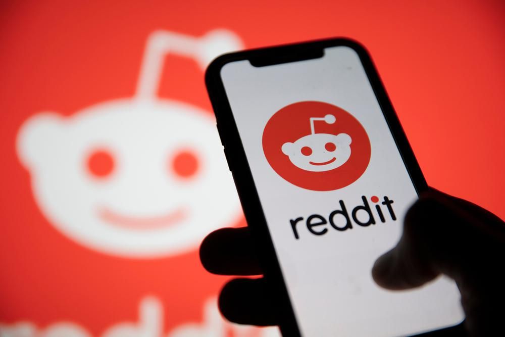 Egy nagy Reddit logó előtt egy ember a kezében tartja az okostelefonját, amin épp a Reddit applikációját nyitja meg