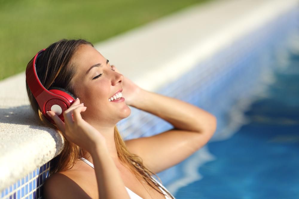 Egy fehér fürdőruhás barna hajú lány verősfényes napsütésben hangoskönyvet hallgat a Spotify-on egy medencében