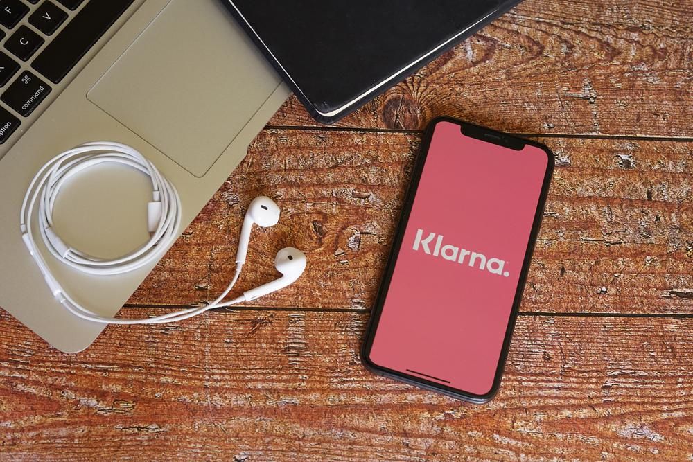 Egy barna faasztalon van egy laptop, egy fülhallgató és egy okostelefon, amin a Klarna applikációja töltődik