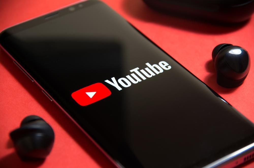 A YouTube applikációja tölt egy okostelefonon, ami mellett két bluetooth fülhallgató található egy piros asztalon
