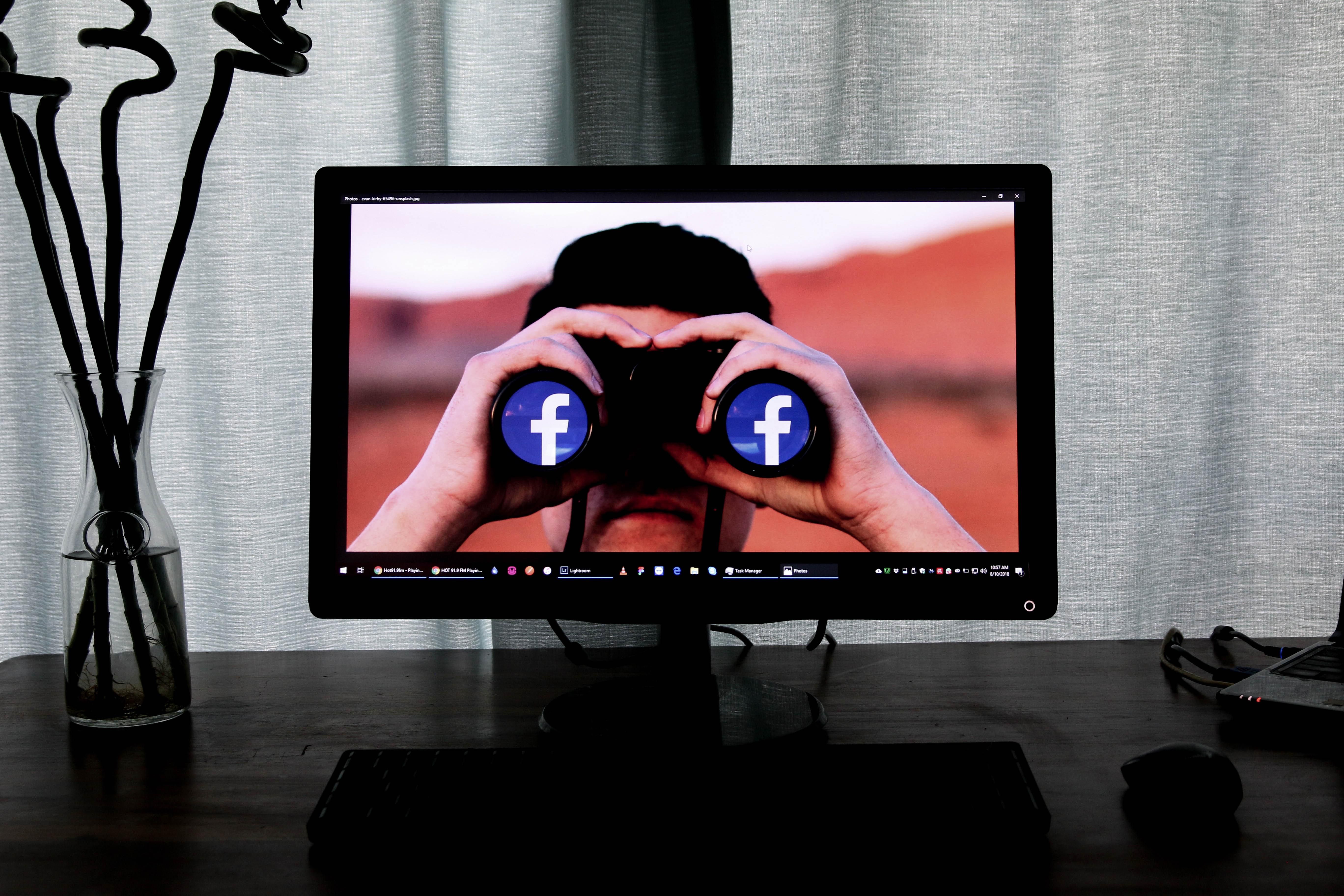 Egy szürke függöny előtt áll egy sötétbarna asztal, amin van egy egér, egy klaviatúra, egy váza és egy monitor, a monitoron egy ember látható, aki Facebook emblémás távcsőbe néz