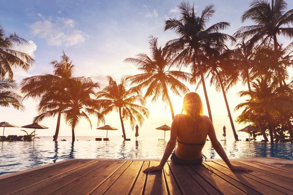 Egy kék fürdőruhás lány az óceán partján egy mólón ülve, pálmafák között, naplementében élvezi az évi 365 fizetett szabadságát