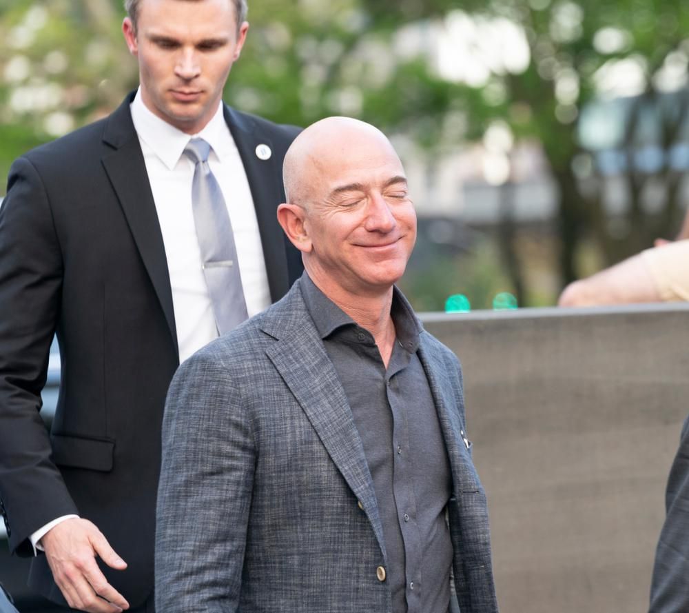 Jeff Bezos szürke öltönyben mosolyog egy fekete zakós ember előtt napsütéses időben, csukott szemmel