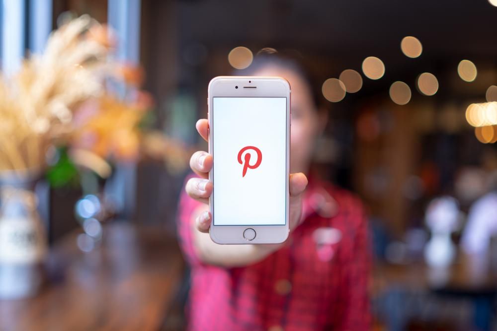 Elmosódott háttérrben egy piros-fekete inges lány a kezében tart egy okostelefont, amelyen a Pinterest logója látható 