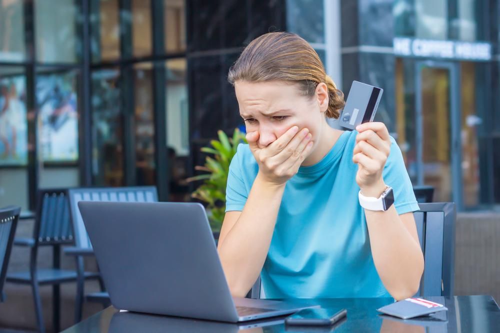Egy kávézó szabadtéri asztalánál ülő kék pólós nő elképedve fogja az arcát kezében a bankkártyájával, mert a laptopját nézve rájött, hogy átverték a csalók