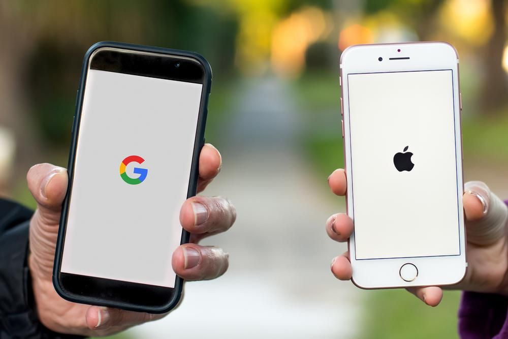 Egy férfi egy Androidos telefont tart a kezében, amelyen a Google logója látható fehér háttérrel, és egy nő egy iPhone-t tart a kezében, amelyen az Apple logója látható fehér háttérben