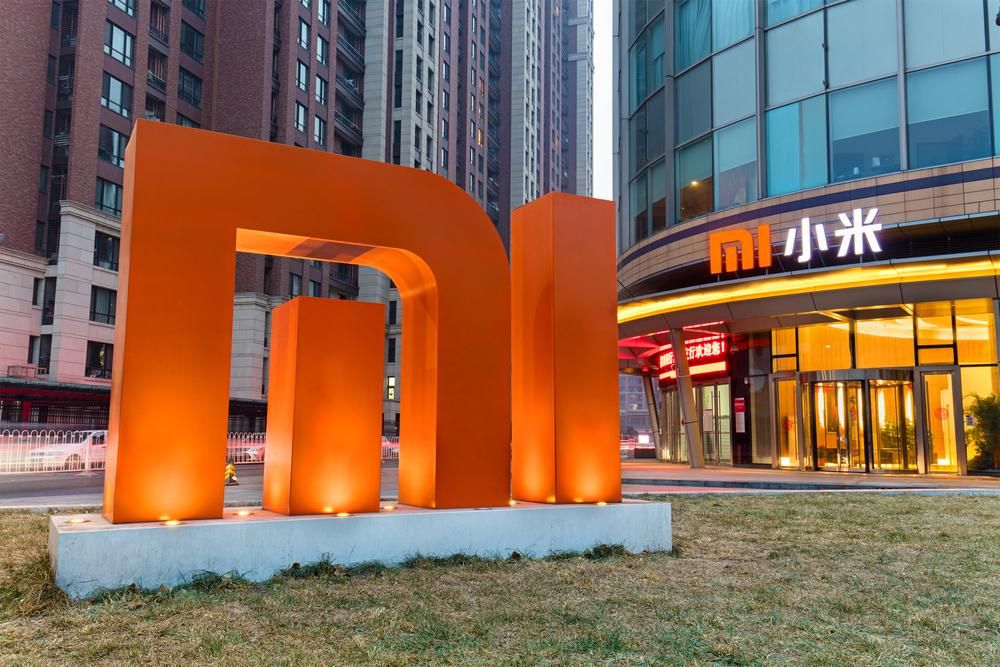 A Xiaomi logójának installációja egy füve téren a Xiaomi egyik üzlete előtt, amelynek homlokzatán látható a Xiaomi logója kínai írásjelek mellett egy forgalmas utcán, lakóépületek között 