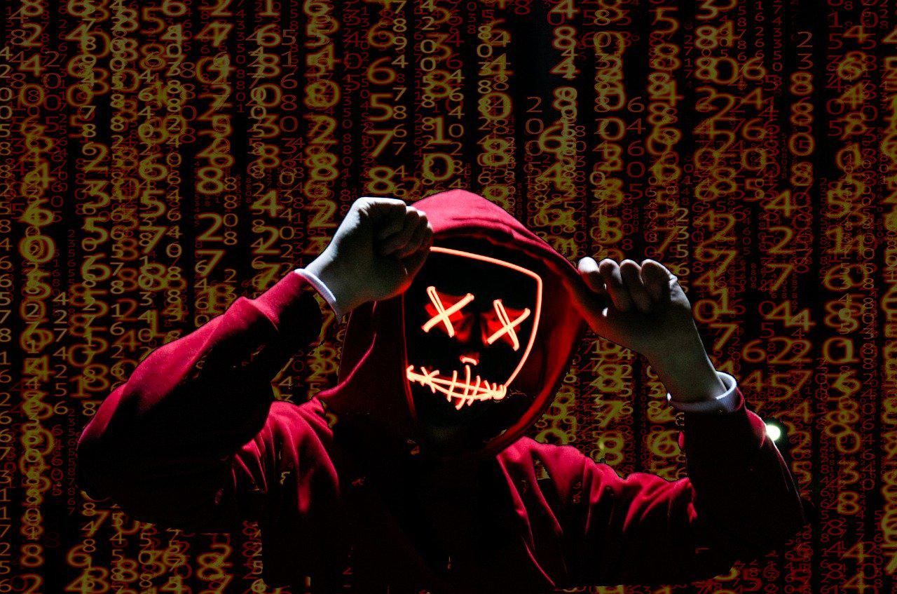 Narancssárga, vertikális számsorok előtt áll egy piros pulcsis hacker, a kapucniját fogja, maszkban van, a szeme két x, a szája össze van varrva