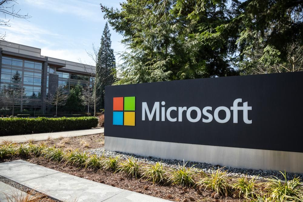 A Microsoft logója egy kavicsos virágágyás mögött és egy fenyőfa előtt a cég irodaközpontjának udvarán