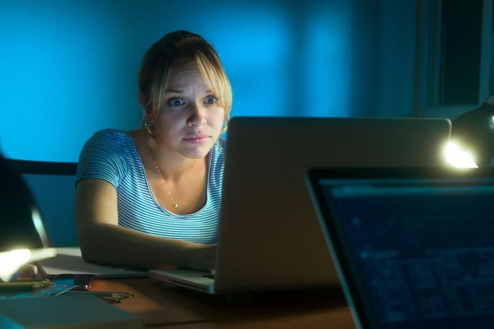 Csíkos pólós szőke nő este meglepődve nézi a laptopja képét, miközben az adóhivatal üzenetét olvassa Facebookon