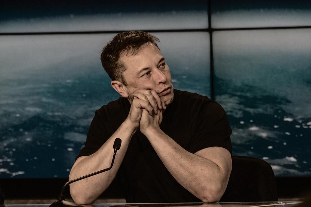 Elon Musk fekete pólóban könyököl  egy asztalon egy mikrofon előtt