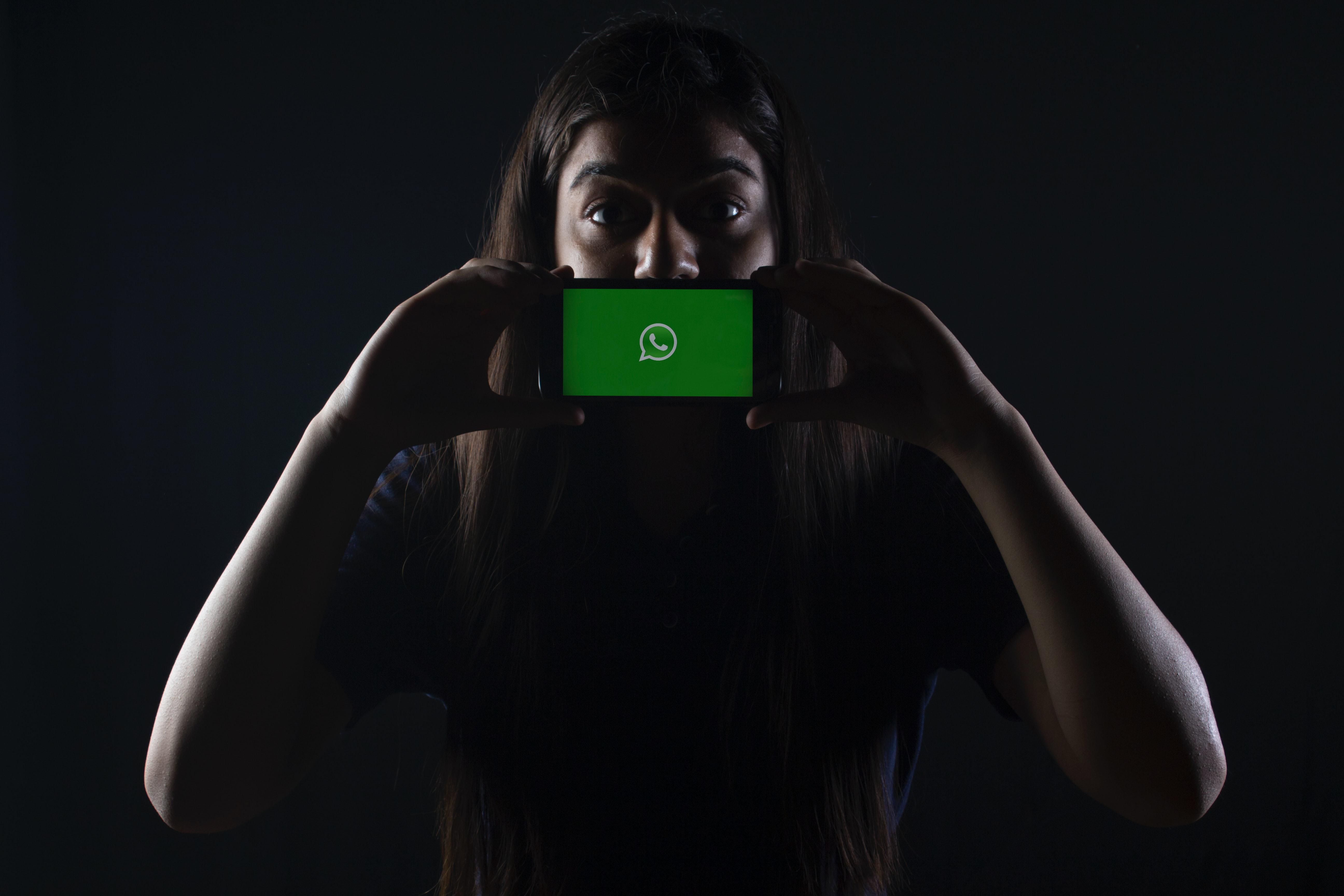Hosszú barna hajú kék pólós lány félhomályban egy okostelefont tart a szája előtt, amin a WhatsApp logója látható